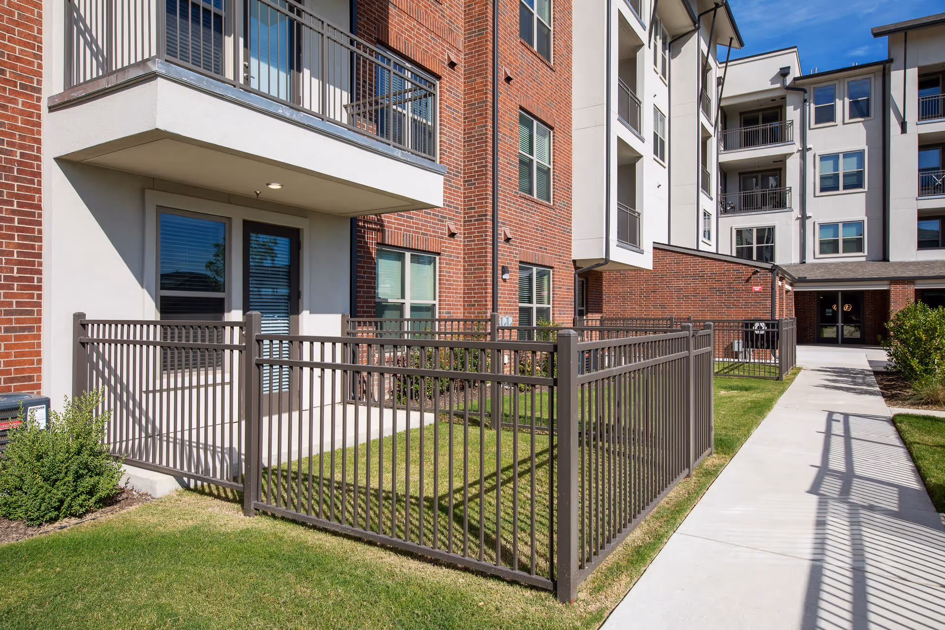 Outdoor view of a senior living facility showing a fenced patio area with green grass, a concrete walkway, and a multi-story building with brick and white exterior walls under a clear blue sky.