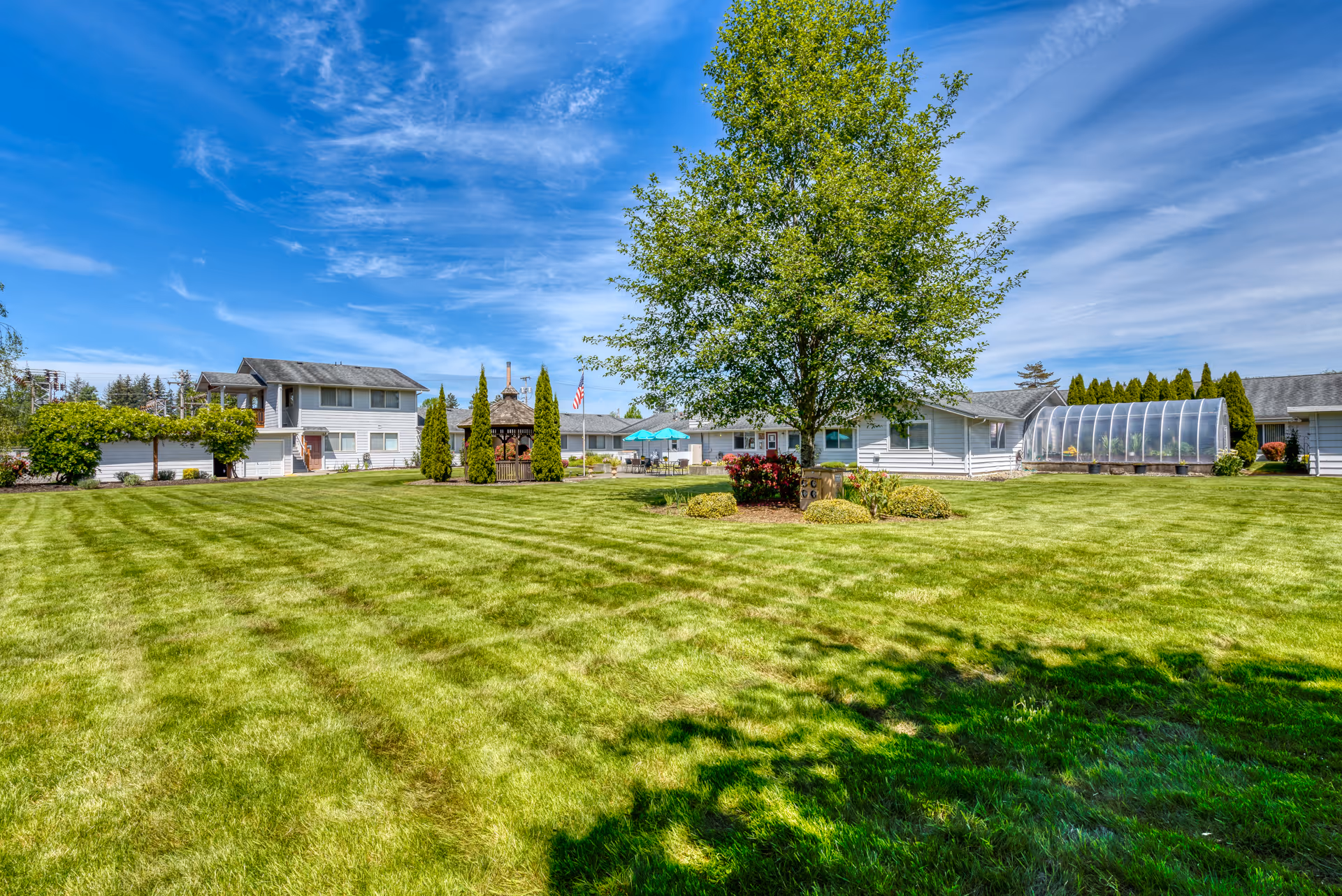 A large, well-maintained grassy lawn with a tree and shrubs in the center, surrounded by single-story and two-story white buildings under a blue sky with scattered clouds. There is a gazebo, patio furniture with umbrellas, and a greenhouse structure visible in the background.