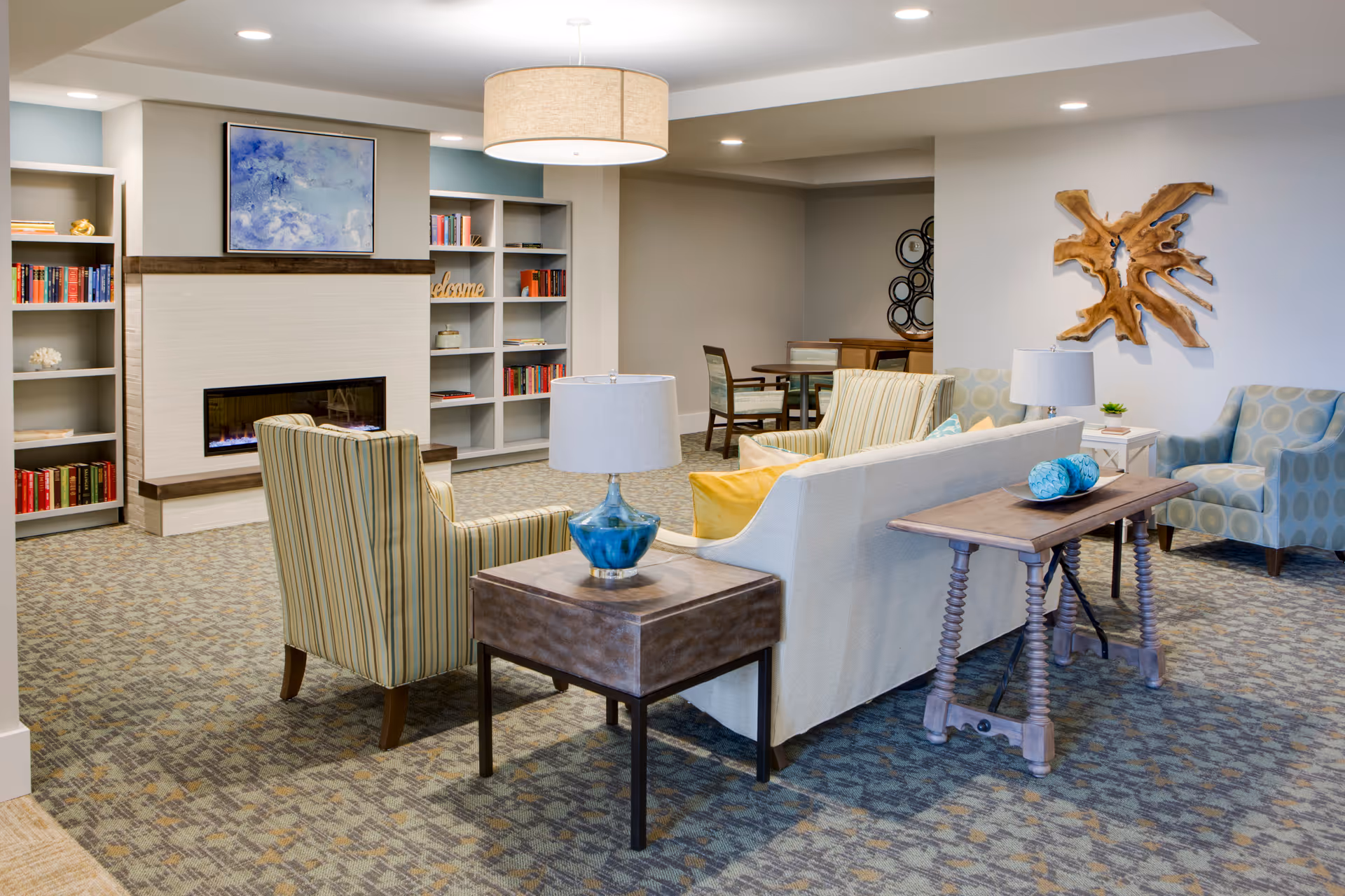 Bright communal living room with sofas and chairs arranged around side tables, a fireplace, built-in bookshelves, and wall decor.