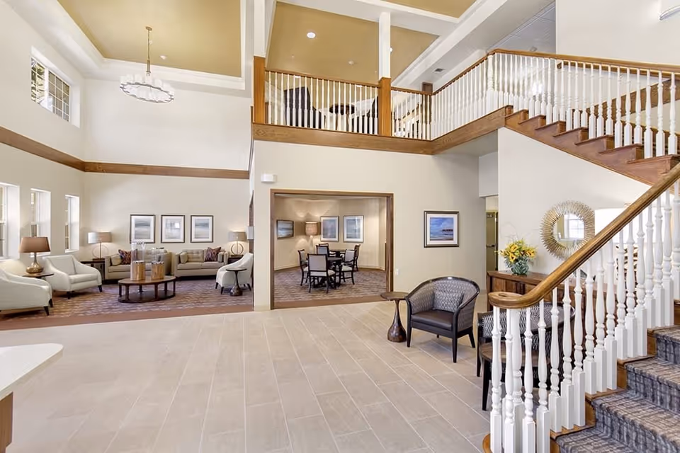 Spacious senior living facility common area with a high ceiling, beige tiled floor, and a wooden staircase with white railings. The room features a seating area with armchairs and sofas around a coffee table, a dining area with a table and chairs, framed artwork on the walls, and decorative lamps. There is a second-floor balcony overlooking the space.