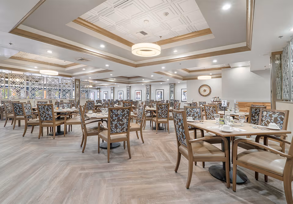 Spacious dining room with multiple wooden tables and chairs featuring floral patterned upholstery. The room has a light wood floor, decorative ceiling with recessed lighting, and large windows allowing natural light. Tables are set with white cups, glasses, and silverware.