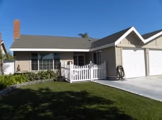 Single-story house with beige siding, a gray shingled roof, a white picket fence enclosing a small front porch, a two-car garage, and a well-maintained green lawn in front.