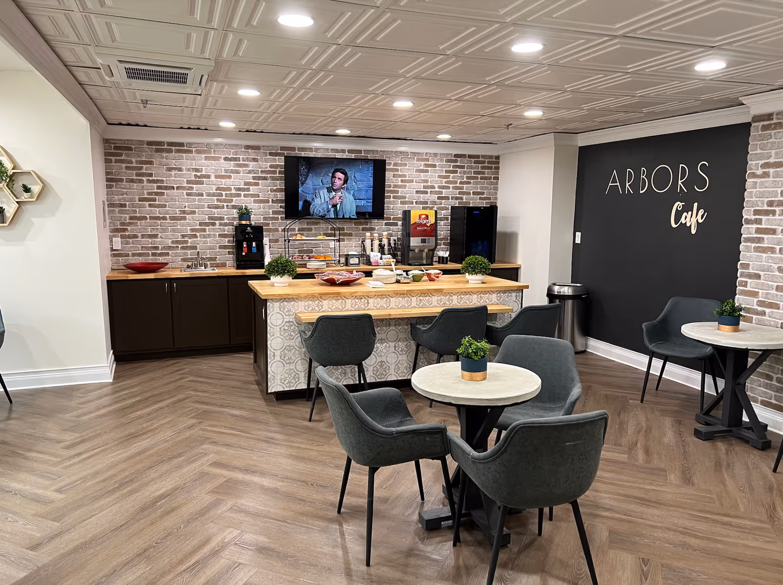 Cozy cafe-style dining area with round tables and chairs, a serving counter against a brick wall, and 'ARBORS Cafe' signage.
