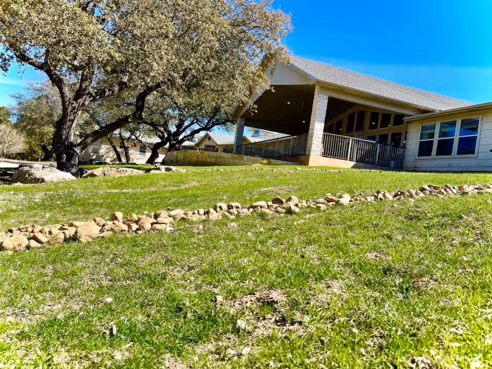 Outdoor view of Heritage Place of Boerne showing a grassy area with a stone-lined path, mature trees, and a building with a covered porch and large windows under a clear blue sky.