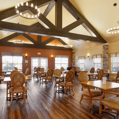 A spacious dining room with wooden floors, multiple wooden tables and upholstered chairs arranged neatly. The room features high vaulted ceilings with exposed dark wooden beams and circular chandeliers. Large windows with blinds allow natural light to fill the room, and the walls are painted in warm tones with stone accents.