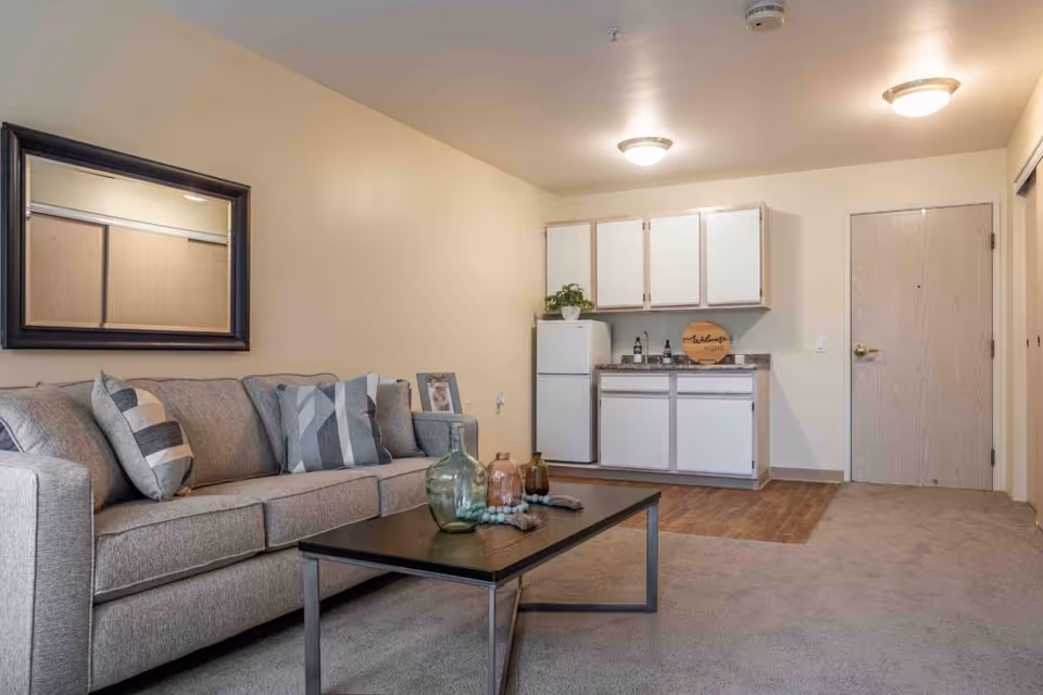 A cozy living area in a senior living facility featuring a gray sofa with patterned cushions, a black coffee table with decorative glass bottles, a wall mirror above the sofa, and a small kitchenette with white cabinets, a refrigerator, and a countertop. The room has beige walls, carpeted flooring, and two ceiling lights.