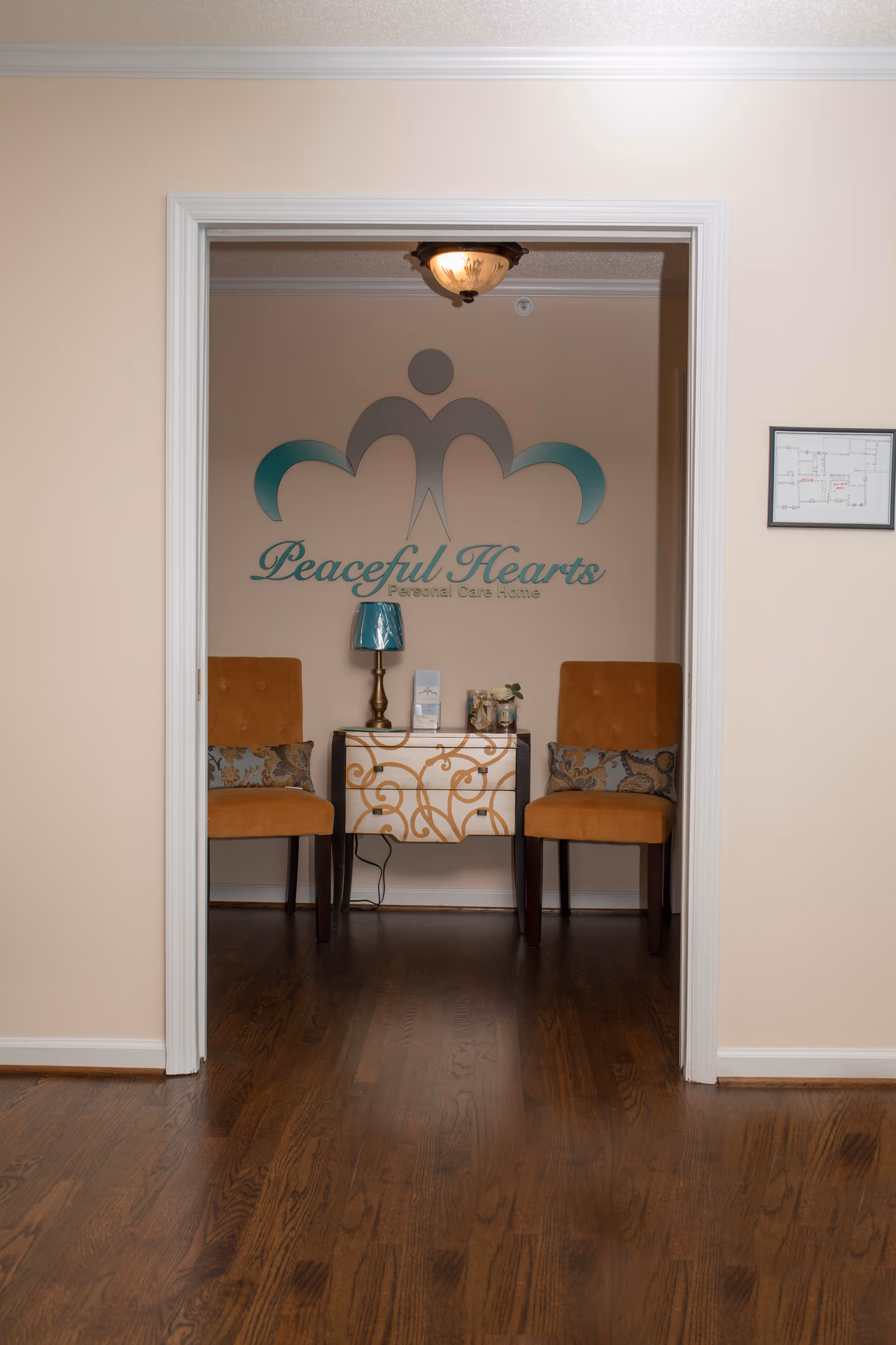 Interior view of a small seating area in Peaceful Hearts Personal Care Home with two orange upholstered chairs on either side of a decorative white and gold table. A small lamp and some decorative items are on the table. The wall behind features the Peaceful Hearts logo and name. The floor is wooden and the walls are beige.