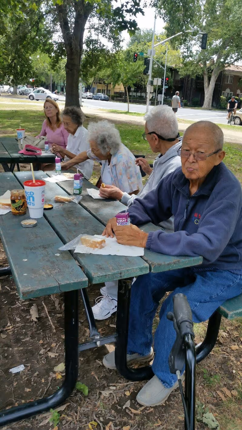 A group of elderly people sitting at a green wooden picnic table outdoors in a park. They are eating snacks and drinking from juice boxes and cups. Trees and a street with cars and traffic lights are visible in the background.