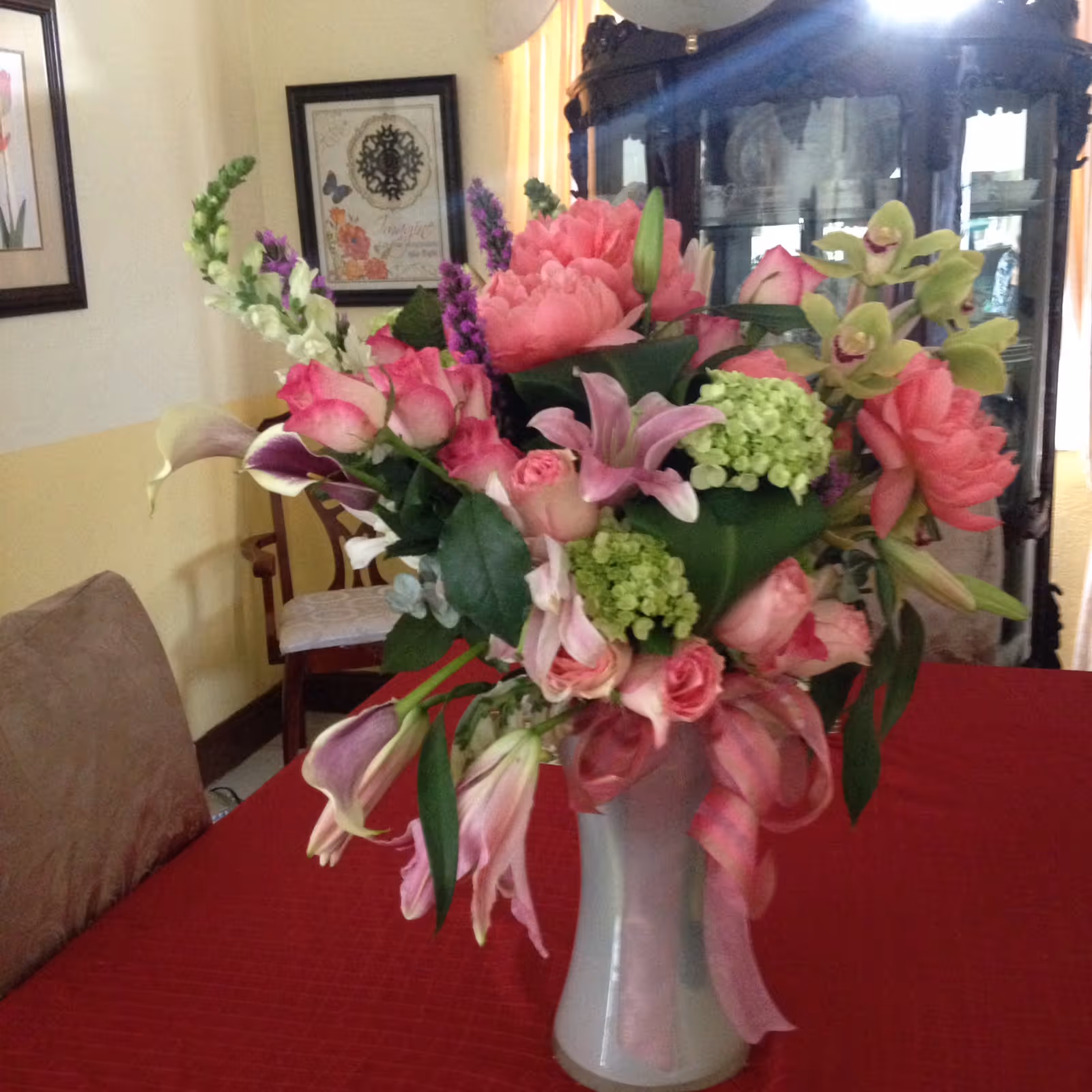 A vase of pink and green flowers arranged on a red tablecloth in a dining room with chairs and a china cabinet in the background.