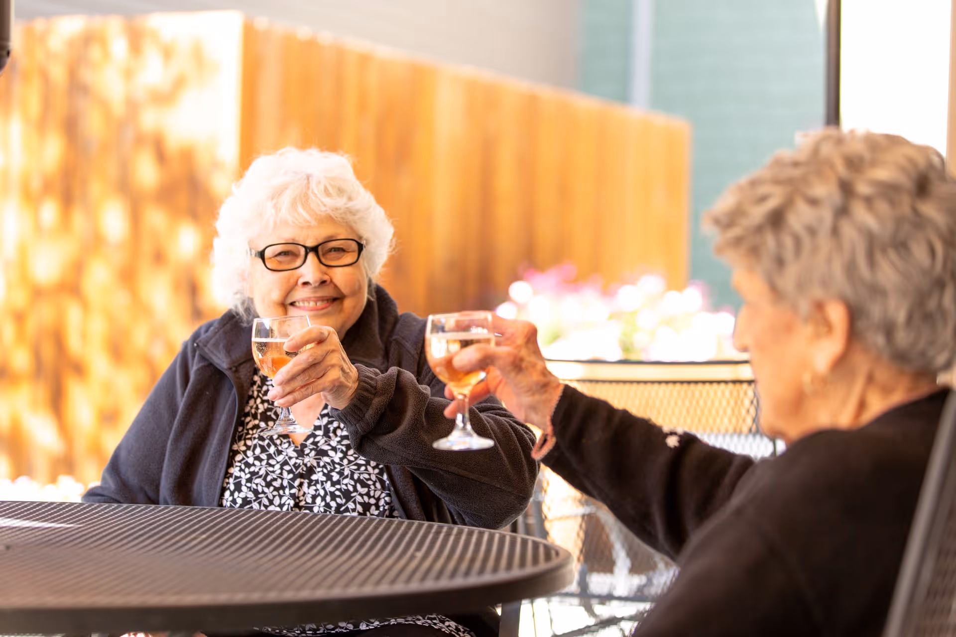 Two elderly women sitting at a round outdoor table, smiling and clinking glasses of a light-colored beverage in a toast. One woman has white hair and glasses, wearing a dark jacket over a patterned blouse. The other woman has short gray hair and is wearing a dark sweater. The background shows a wooden fence and some blurred flowers.
