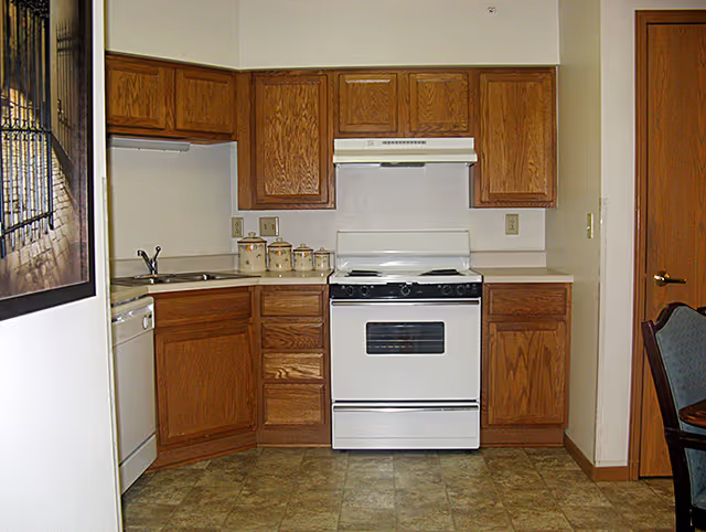 A small kitchen with wooden cabinets, a white electric stove with oven, a white dishwasher, and a countertop with a set of canisters. The floor has a tile pattern, and there is a partial view of a dining chair on the right side.