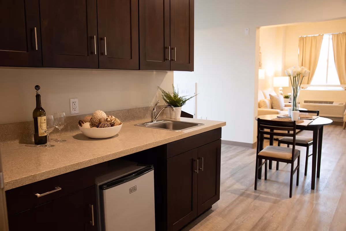 A kitchenette area with dark wood cabinets, a small sink, a mini refrigerator, and a countertop decorated with a bottle of wine, two wine glasses, a bowl of decorative balls, and a small potted plant. In the background, there is a dining table with four chairs and a vase with white flowers, leading to a living area with a sofa, lamps, and a window with beige curtains.