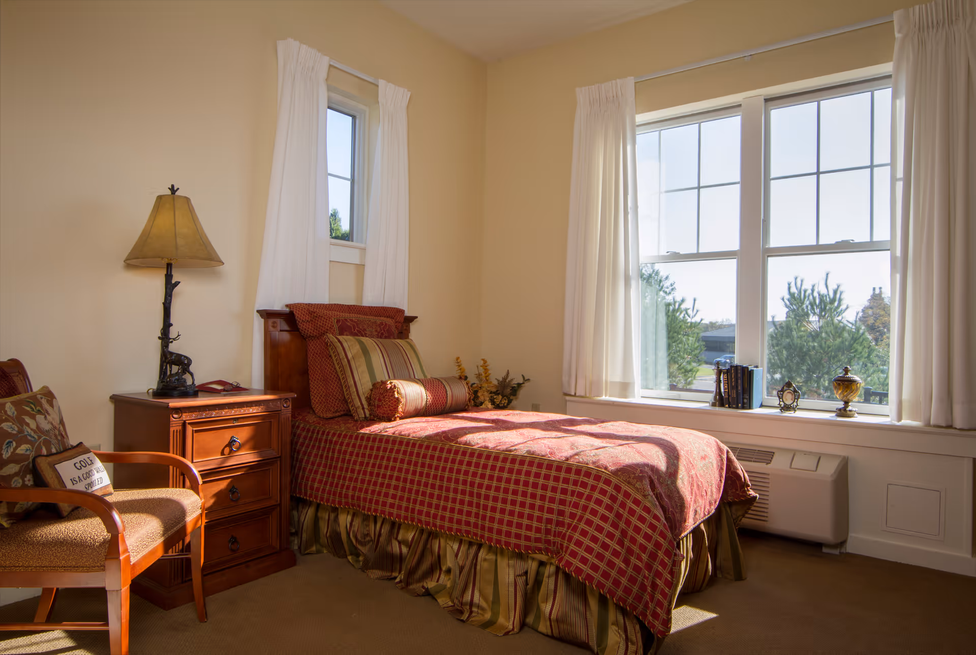 Sunlit bedroom with a single bed, wooden nightstand and lamp, a chair, and large windows.