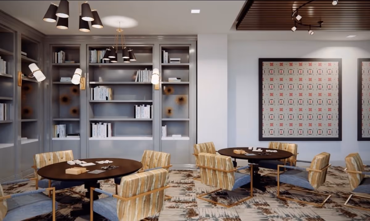 A modern common area with two round tables surrounded by cushioned chairs with wooden armrests. The room features built-in bookshelves filled with books, contemporary wall sconces, and pendant lights hanging from the ceiling. The floor has a patterned carpet, and there are framed artworks on the white walls.