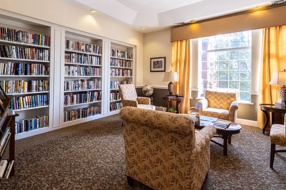A cozy library room with built-in bookshelves filled with books along one wall. The room features patterned armchairs and a sofa arranged around a wooden coffee table. Large windows with yellow curtains allow natural light to brighten the space, and two table lamps provide additional lighting.