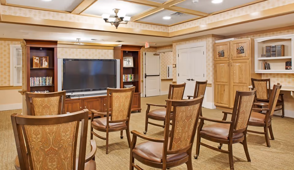 A cozy common room with multiple wooden chairs arranged facing a large flat-screen TV mounted on a wooden entertainment center. The room has beige patterned wallpaper, carpeted floor, and built-in wooden cabinets and shelves with books and framed photos. The ceiling features recessed lighting and a chandelier.