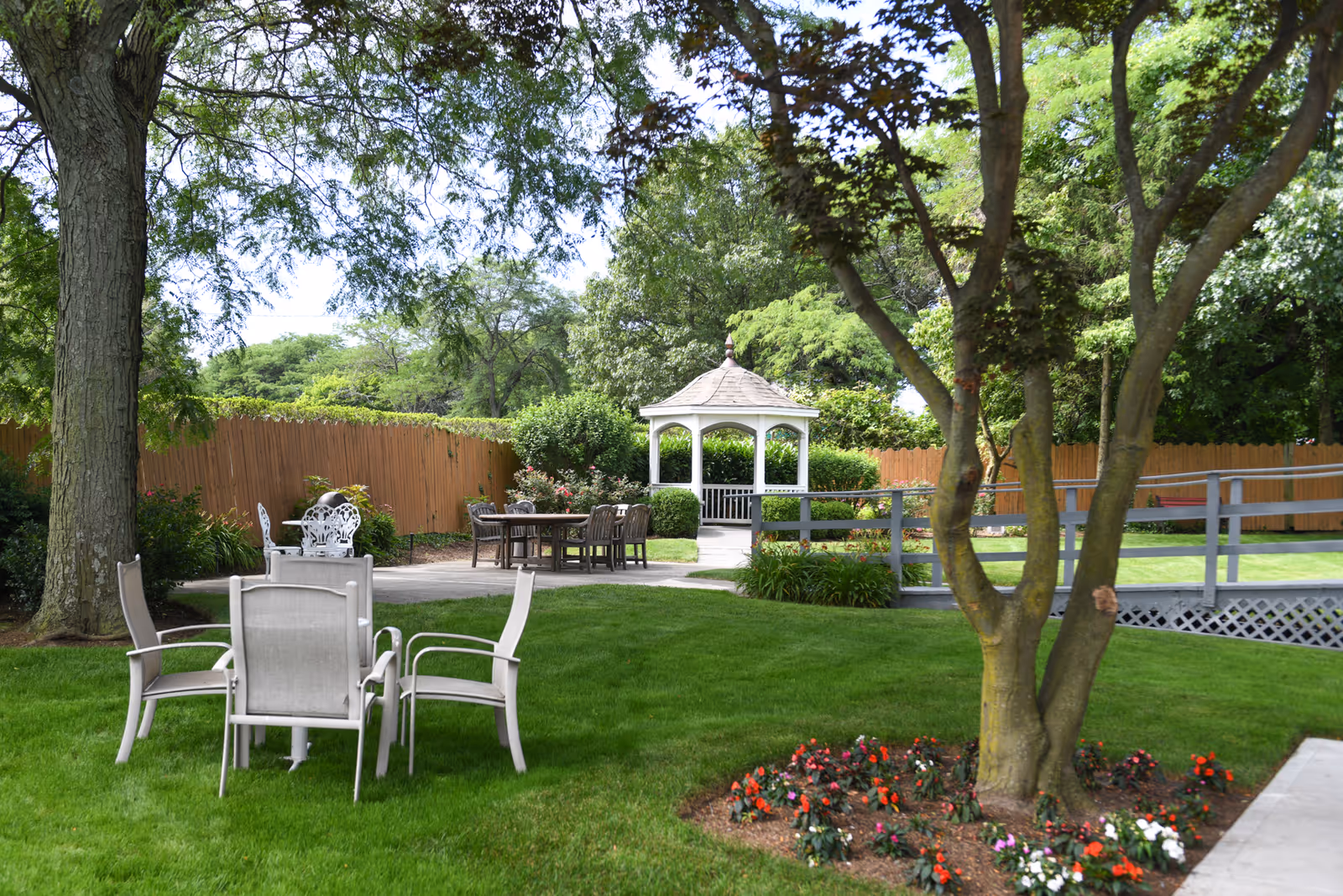 A peaceful outdoor garden area with green grass, trees, and flower beds. There are several seating arrangements including a set of four chairs around a small table in the foreground, and another table with chairs further back near a white gazebo. A wooden fence encloses the garden space.