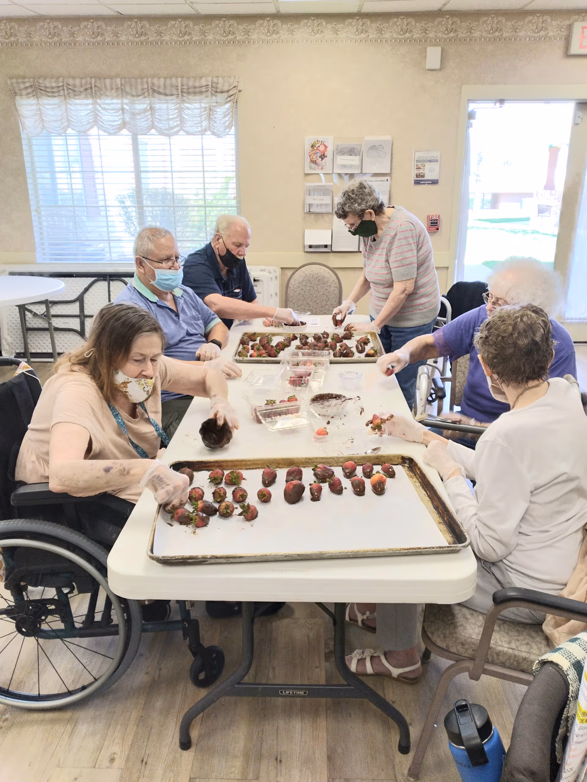 A group of elderly individuals sitting around a table in a well-lit room, dipping strawberries in chocolate and placing them on trays. Some are wearing masks and gloves, and the room has a large window with blinds and a door leading outside.