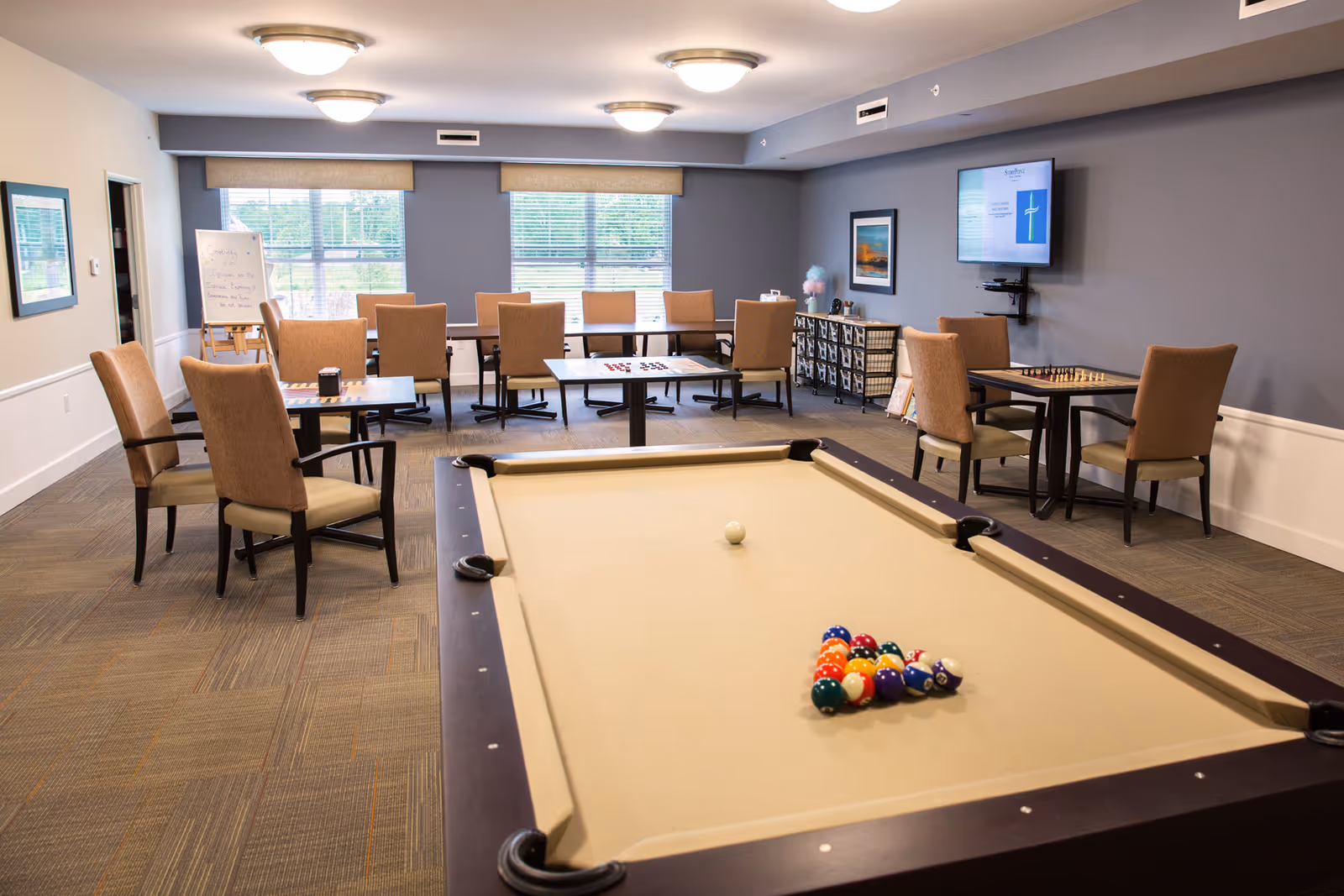 A recreational room with a beige pool table in the foreground, set up with billiard balls and a cue ball. In the background, there are several tables with chairs arranged around them, some with board games like chess and checkers. The room has large windows with blinds, a wall-mounted TV, and framed artwork on the walls. The carpet is patterned, and the lighting is provided by ceiling fixtures.