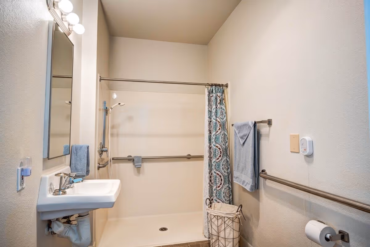 A clean and accessible bathroom featuring a walk-in shower with grab bars, a patterned shower curtain, a wall-mounted sink with a mirror above it, blue towels hanging on towel racks, a laundry basket, and a toilet paper holder.