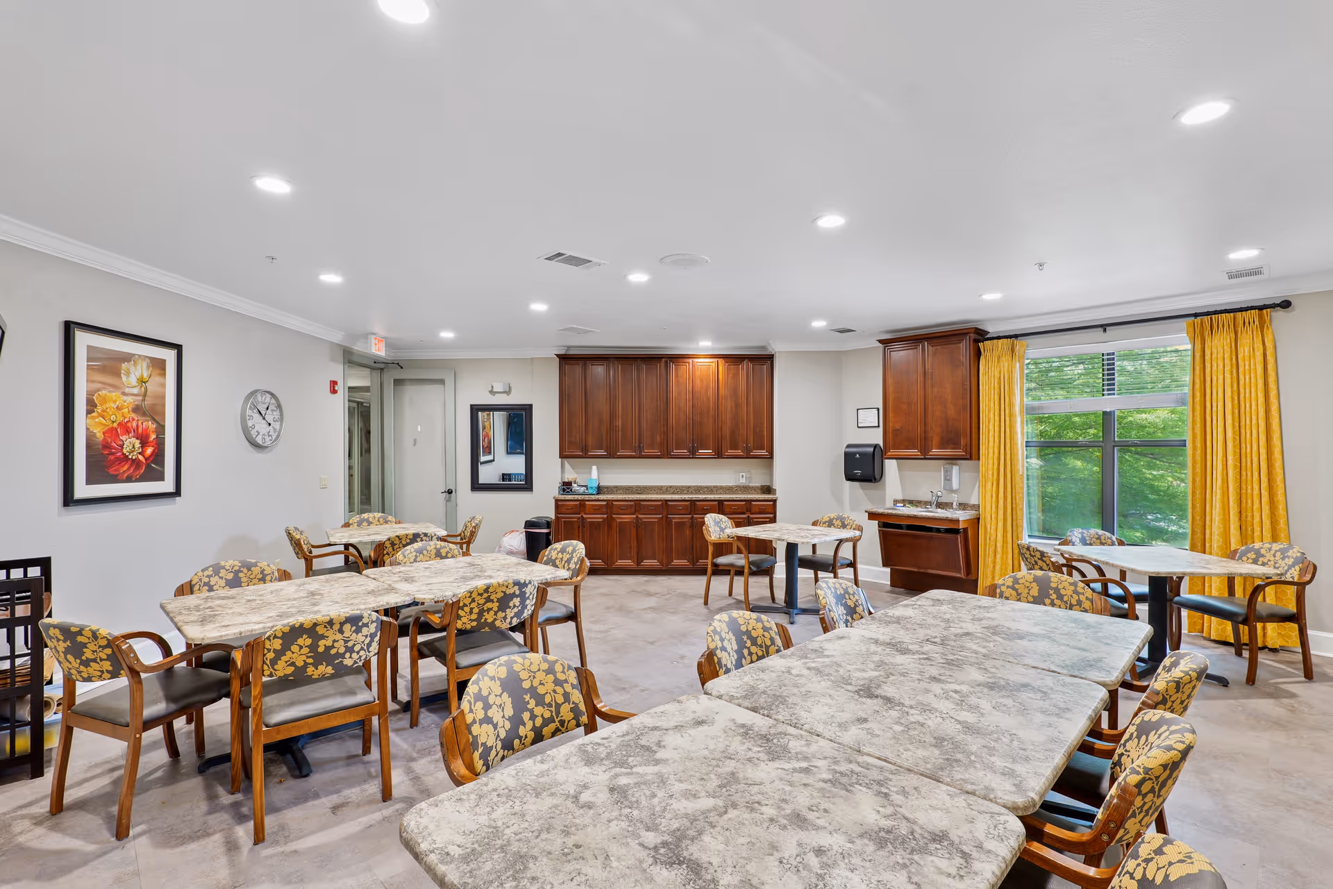 A bright dining room with multiple tables and chairs featuring floral upholstery. The room has a large window with yellow curtains, wooden cabinets along the back wall, a clock, and framed floral artwork on the walls. The floor is tiled and the ceiling has recessed lighting.