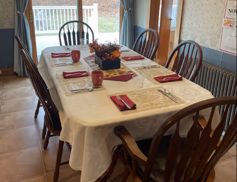 A dining table set for six with a cream-colored tablecloth, red napkins, silverware, clear drinking glasses, and red mugs. A centerpiece with autumn-themed flowers is in the middle of the table. The room has wooden chairs around the table, a window with blue curtains, and a radiator along the wall.