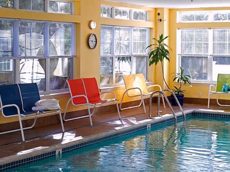 Indoor pool area at Atria Lincoln Place featuring colorful lounge chairs, large windows, and a tropical plant.