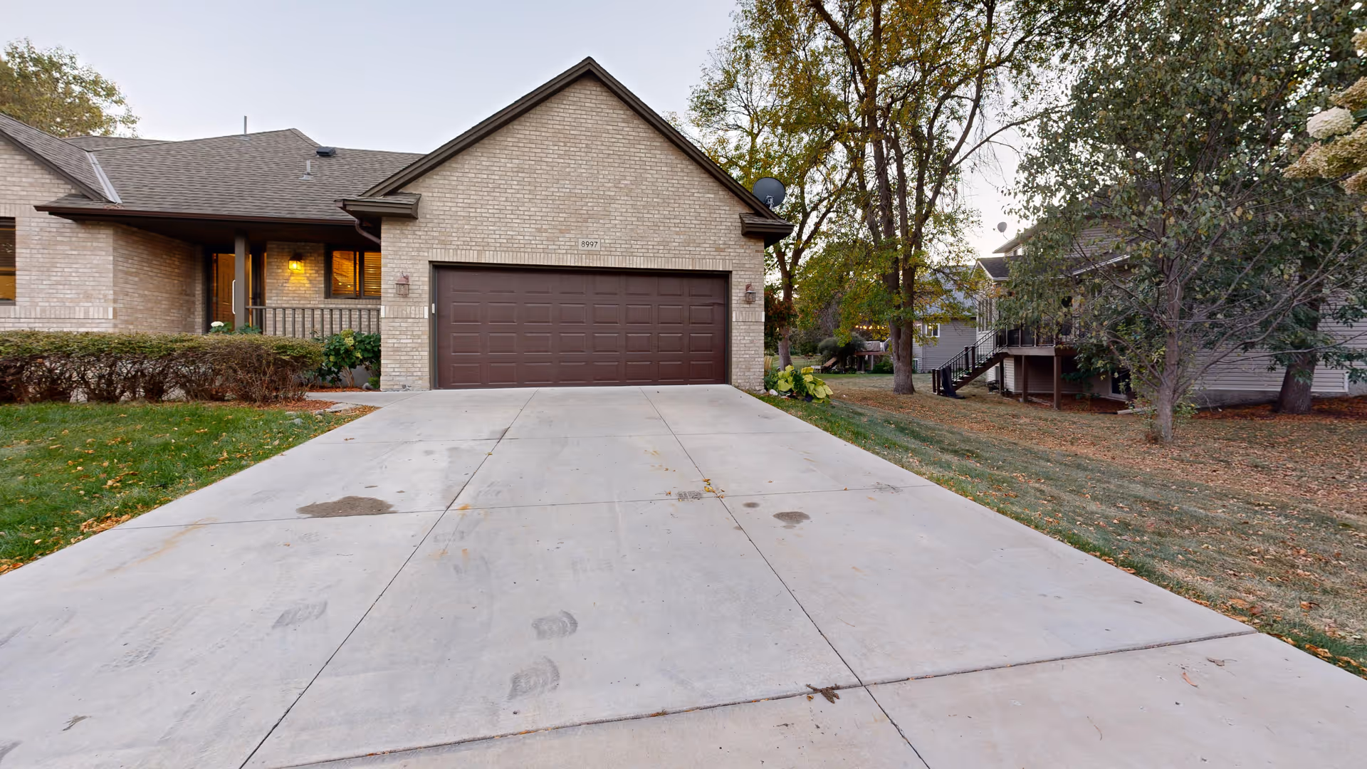 Front exterior view of a single-story brick house with a large concrete driveway leading to a double garage door. There are trees and grass on either side of the driveway, and the house has a covered porch with a light on.