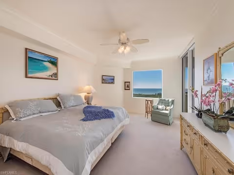 A bright bedroom with a large bed covered in gray and white bedding, a ceiling fan with lights, a green armchair near a window with a view of the ocean, a wooden dresser with a mirror and pink orchids, and framed artwork on the walls.