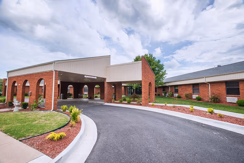 Exterior view of a single-story brick building with a covered entrance driveway, landscaped with grass, shrubs, and mulch beds under a partly cloudy sky.