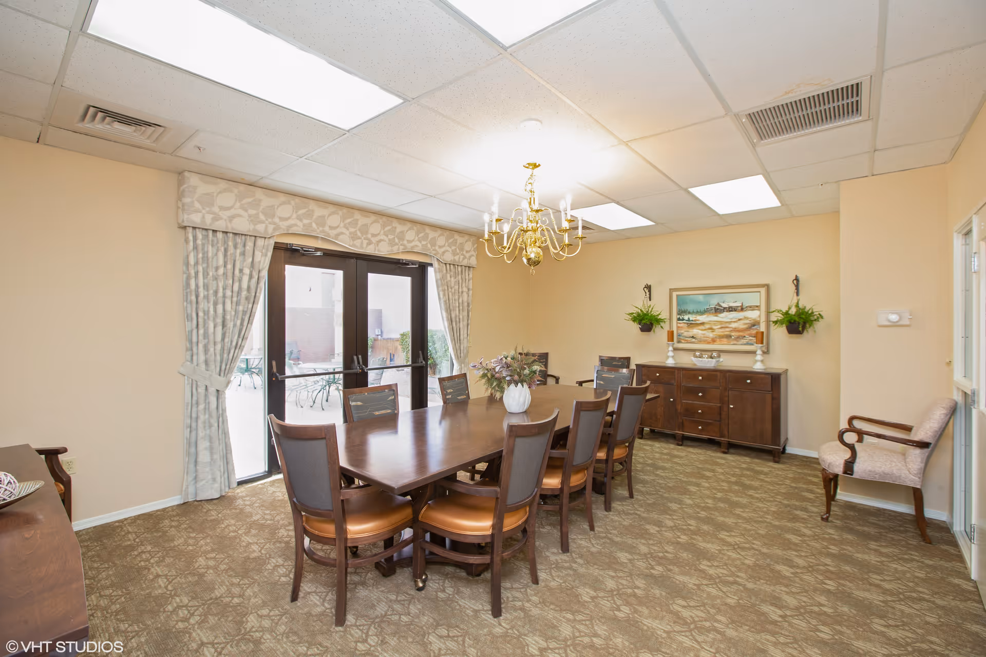 A dining room with a long wooden table surrounded by eight chairs with brown cushions. A white vase with flowers is placed in the center of the table. The room has beige walls, patterned carpet, and a gold chandelier hanging from the ceiling. There are double glass doors with patterned curtains leading outside to a patio area. A wooden sideboard with decorative items and a framed painting is against one wall, with two wall-mounted plants on either side. A single upholstered chair is positioned near the right wall.