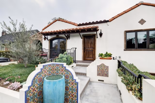Spanish-style white stucco house front with a tiled roof, decorative tiled fountain, steps and a wooden entry door.