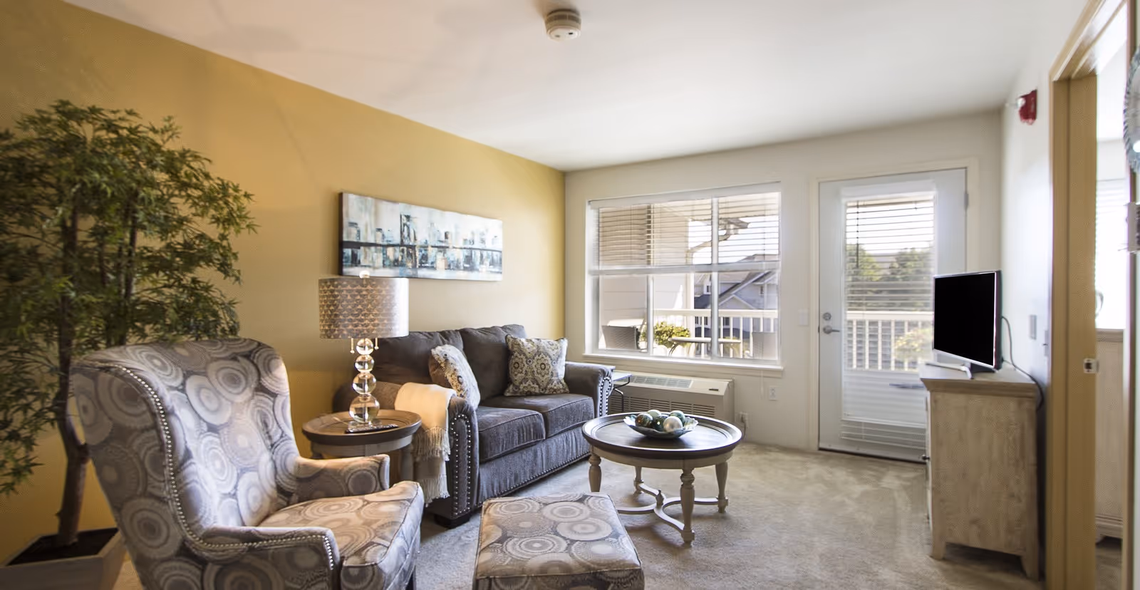 A cozy living room with a patterned armchair and matching ottoman, a gray sofa with decorative pillows, a round wooden coffee table with a bowl of decorative balls, a side table with a lamp, a large potted plant, and a window and door leading to an outdoor balcony.
