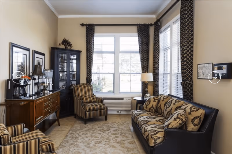 A cozy living room with beige walls and large windows covered with patterned dark curtains. The room features a patterned sofa with matching cushions, two striped armchairs, a wooden sideboard with a coffee maker and other appliances, a black glass-front cabinet, a floor lamp, and a beige patterned rug on the tiled floor.