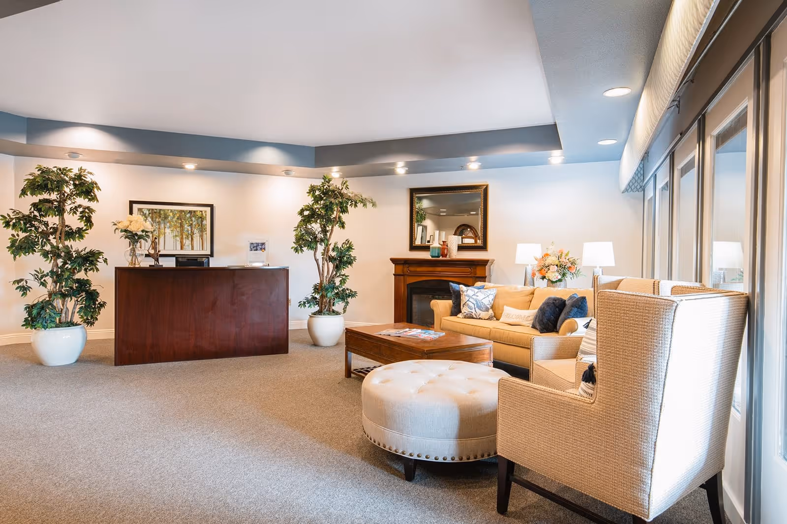 A bright and welcoming reception area with a wooden front desk flanked by two large potted plants. To the right, there is a cozy seating area featuring a beige sofa with decorative pillows, a cushioned ottoman, a wooden coffee table with magazines, and a beige armchair. The room has carpeted flooring, a fireplace with a mirror above it, and large windows letting in natural light.