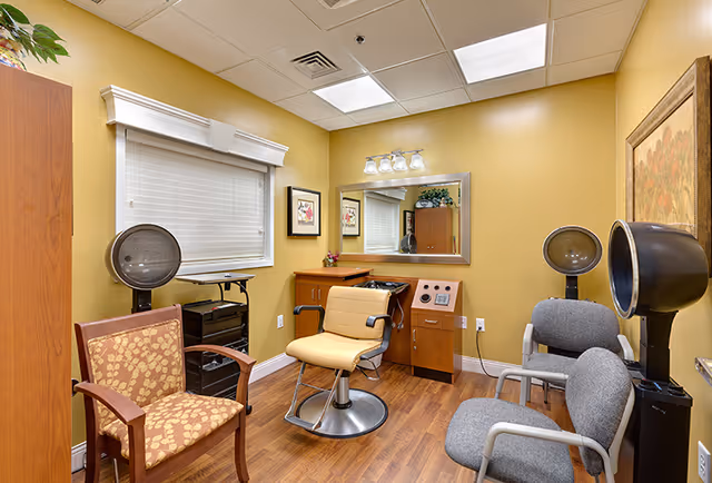 A small salon room with mustard yellow walls and wood flooring. The room contains a central salon chair with a round metal base, two gray chairs with armrests, and a wooden chair with patterned upholstery. There are two black hair dryer stations, a wooden cabinet with a mirror above it, framed artwork on the walls, and a window with white blinds.