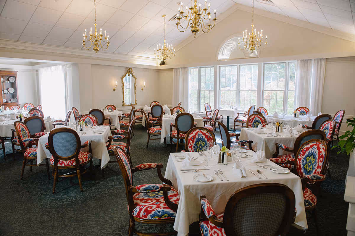 A bright and elegant dining room with multiple tables covered in white tablecloths, set with plates, glasses, and silverware. The chairs have colorful patterned upholstery with red, blue, and yellow designs. The room features large windows allowing natural light, gold chandeliers hanging from a vaulted ceiling, and a decorative mirror on the wall.