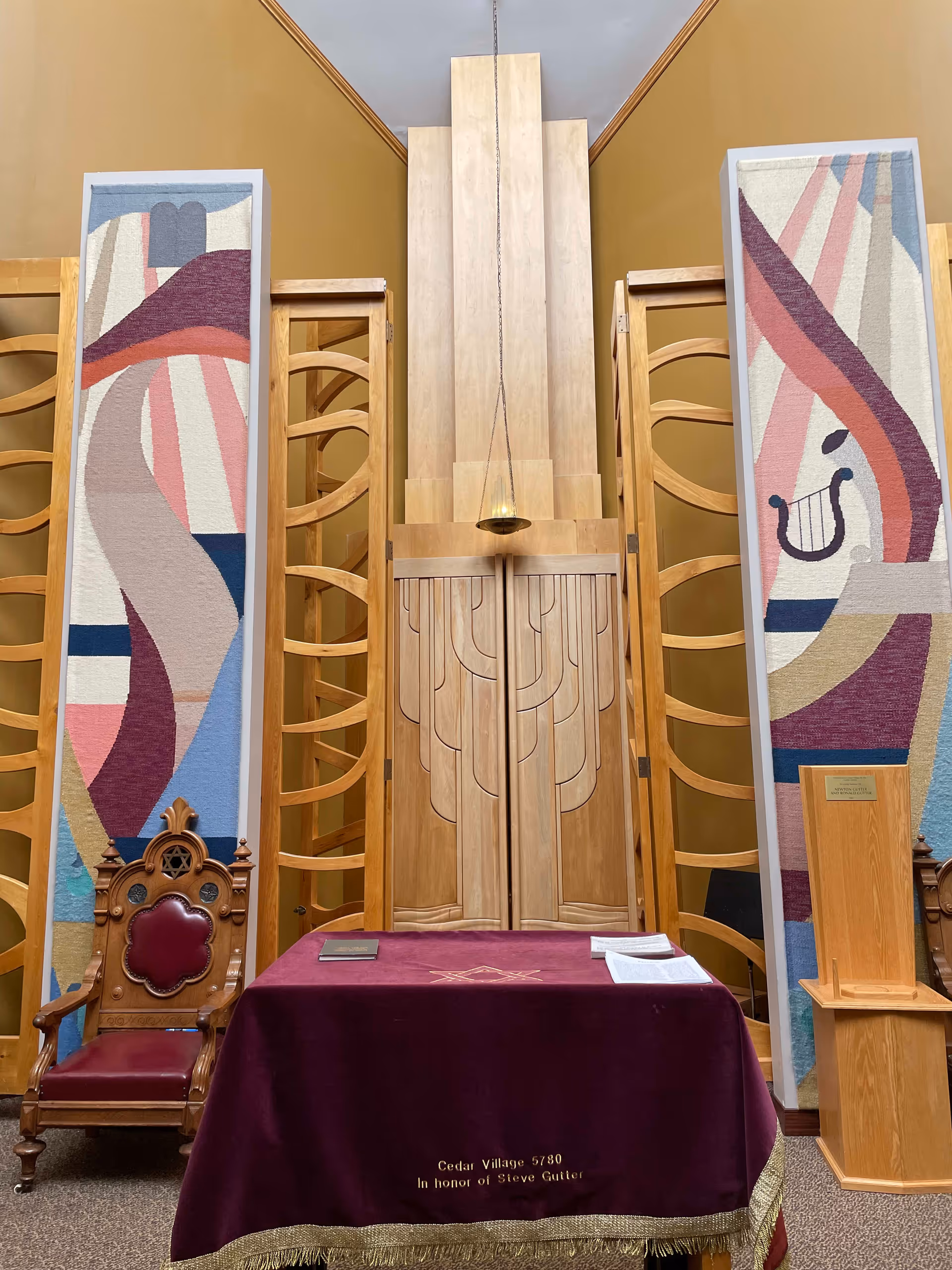 Interior view of a ceremonial or religious room with a wooden chair featuring a Star of David, a table covered with a purple cloth embroidered with a Masonic symbol and the text 'Cedar Village 5780 In honor of Steve Gutter', two tall colorful abstract tapestries on either side, and a wooden cabinet with a hanging lamp above it.