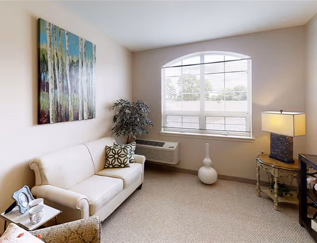 Small bright living room with a loveseat, armchair, side tables, lamp, artwork and a large arched window.