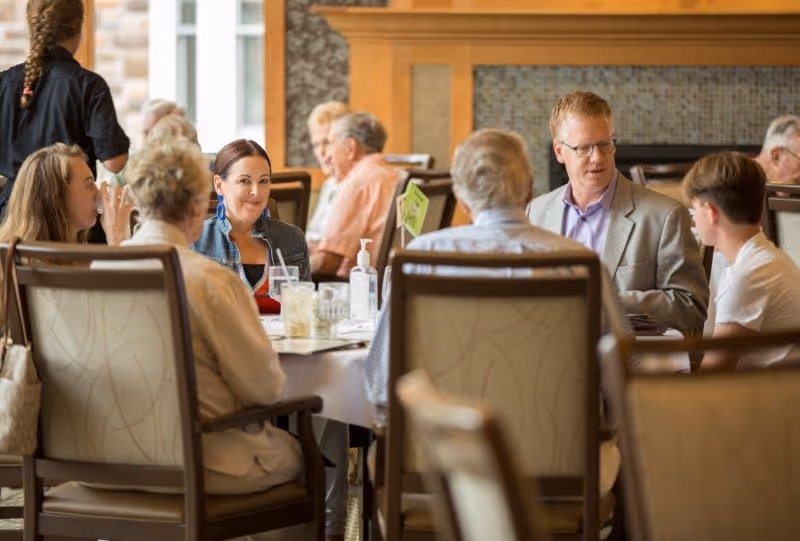 A group of people sitting around a dining table in a well-lit room with a fireplace in the background. They appear to be engaged in conversation and enjoying drinks. The setting looks like a communal dining area in a senior living facility.