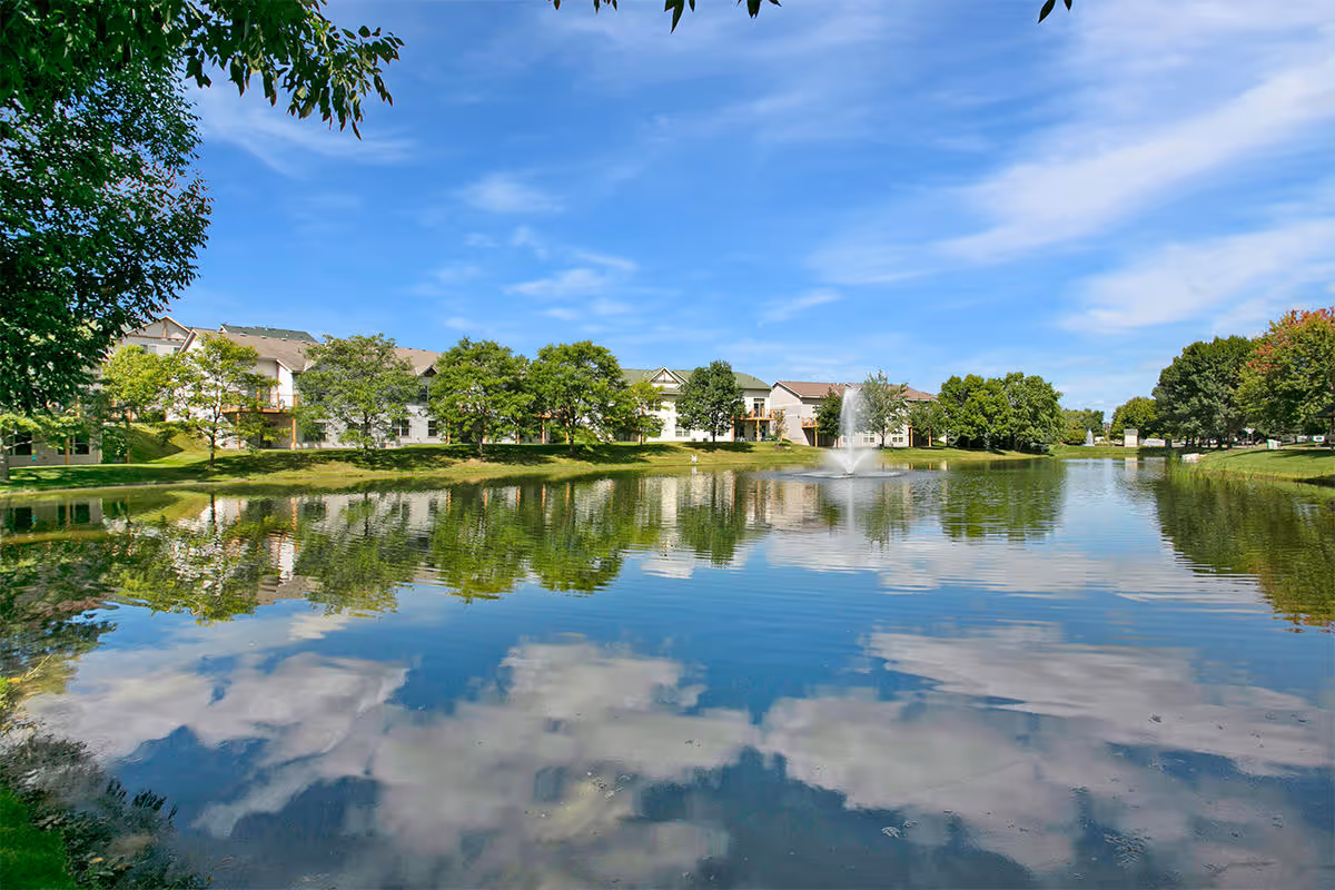 A serene pond with a fountain in the center, surrounded by green trees and residential buildings under a blue sky with scattered clouds. The water reflects the trees, buildings, and sky.