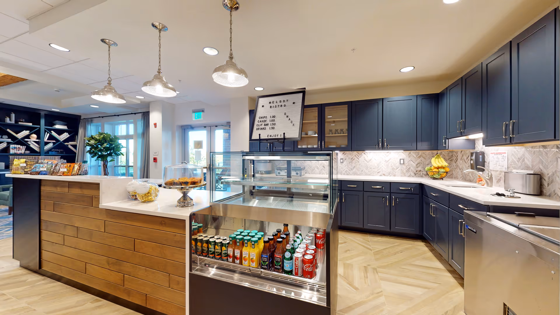 Interior view of a modern snack bistro area in an assisted living facility. The space features a counter with a glass display case containing various bottled and canned beverages. Behind the counter are dark blue cabinets with a white countertop and a sink. Above the counter, three pendant lights hang from the ceiling. A sign on the counter reads 'Melody Bistro' with a list of snack items and prices. In the background, there is a seating area with a bookshelf and large windows letting in natural light.