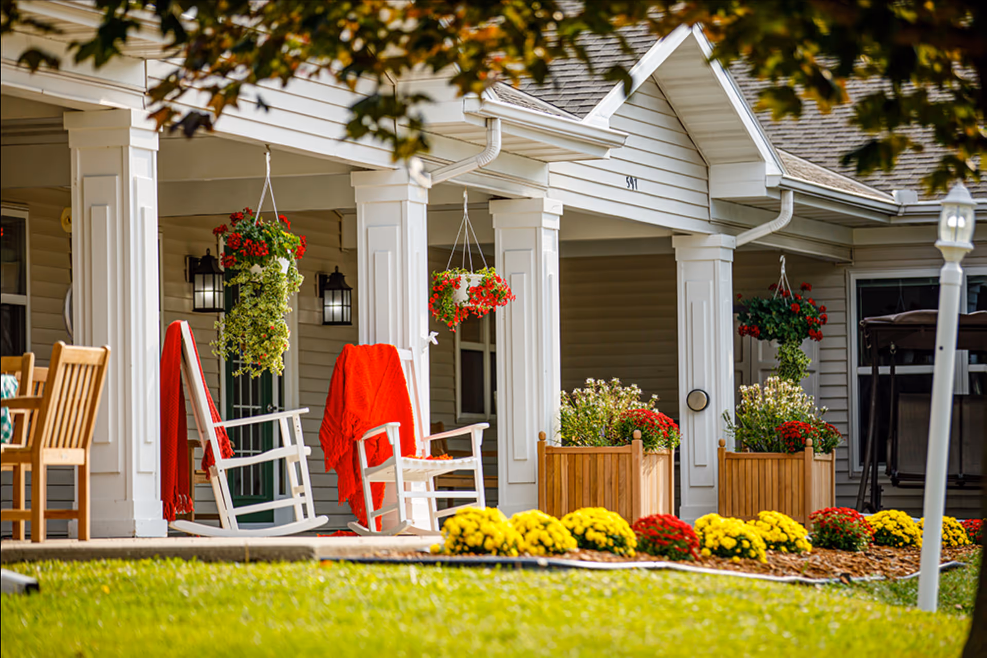 Front porch of a residential building with white columns, two white rocking chairs draped with red blankets, hanging flower pots with red flowers, wooden planters with green and red plants, and a garden bed with yellow and red flowers in the foreground.