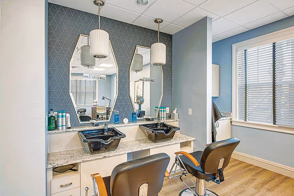 Interior view of a salon area with two black salon chairs in front of a counter with two black wash basins. Above the counter are two uniquely shaped mirrors mounted on a blue wall with a geometric pattern. Various hair care products are placed on the counter. A window with blinds is visible on the right side, allowing natural light into the room.