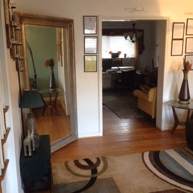 Interior view of a living area in an assisted living facility showing a large ornate mirror leaning against the wall, a small table with a lamp and decorative items, a patterned rug on a wooden floor, and an adjacent room with a desk, chair, and a yellow armchair near a window with blinds.