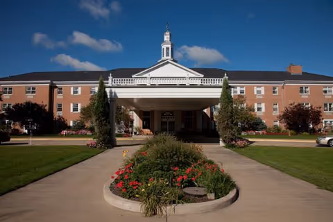 Front exterior view of Copeland Oaks facility, a large brick building with a white portico entrance, manicured lawn, flower beds, and a clear blue sky with some clouds.