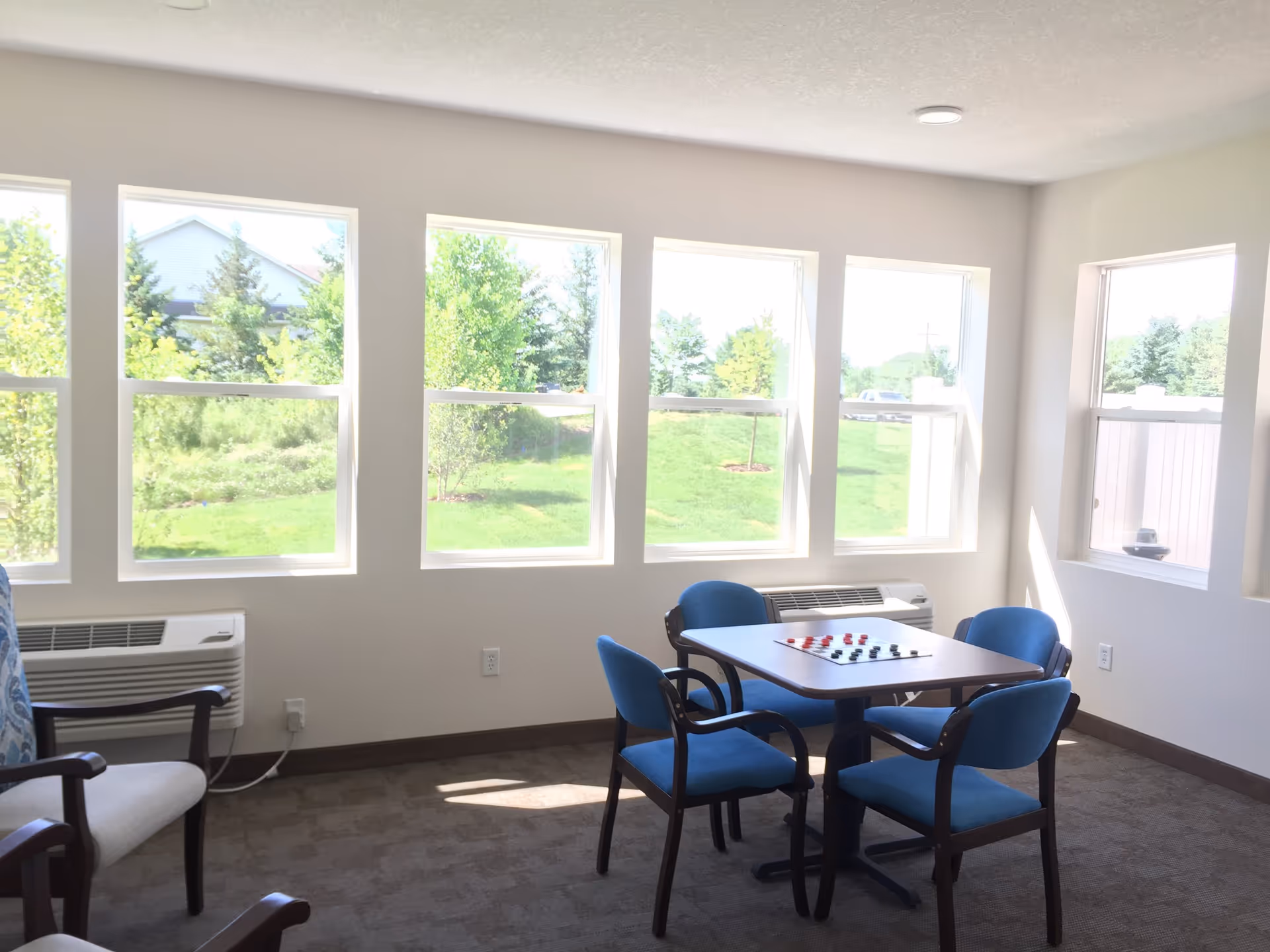 A bright room with five large windows showing a green outdoor area. Inside, there is a square table with a checkers game set up and four blue cushioned chairs around it. There are also two beige cushioned armchairs with wooden armrests partially visible. The room has beige carpet and white walls.