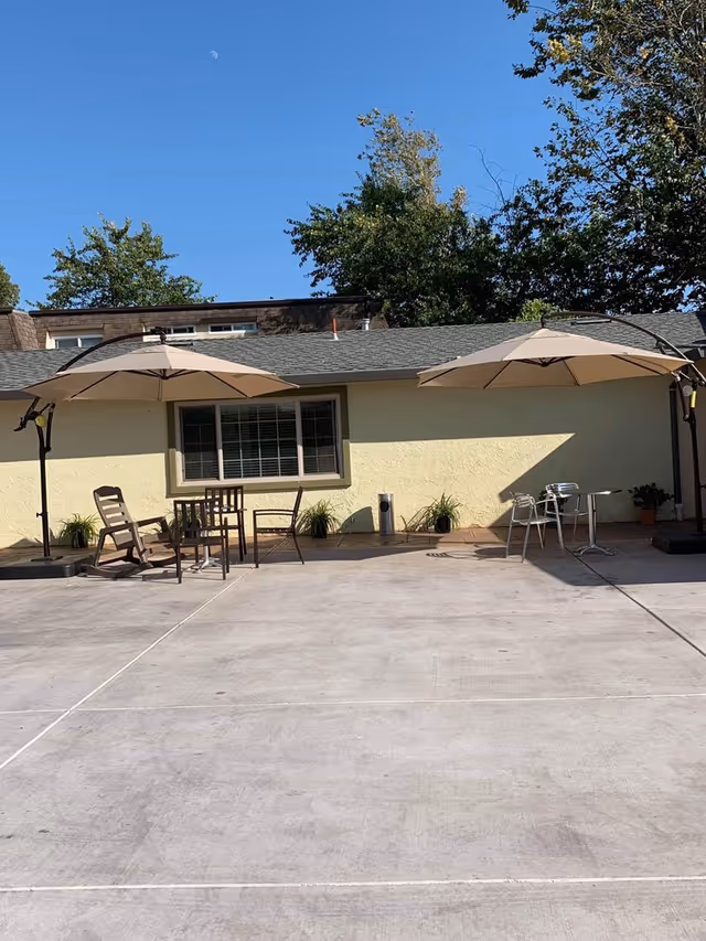 Outdoor patio area with two large beige umbrellas providing shade over several chairs and tables. The patio is adjacent to a light yellow building with a window and some potted plants along the wall. Trees and a clear blue sky with a visible moon are in the background.