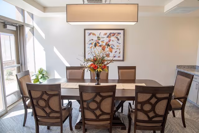 A well-lit dining room with a rectangular wooden table surrounded by eight upholstered chairs. A floral centerpiece is placed in the middle of the table. There is a large rectangular light fixture hanging above the table, a framed artwork on the wall, and a glass door letting in natural light on the left side.