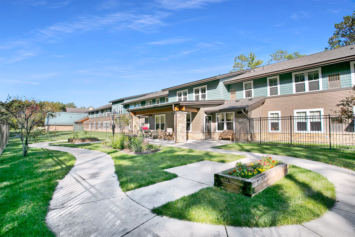 Outdoor view of a senior living facility named Heartwood showing a two-story building with green and brown siding, a covered patio area with seating, a curved concrete walkway, green grass, trees, and a raised flower bed under a clear blue sky.